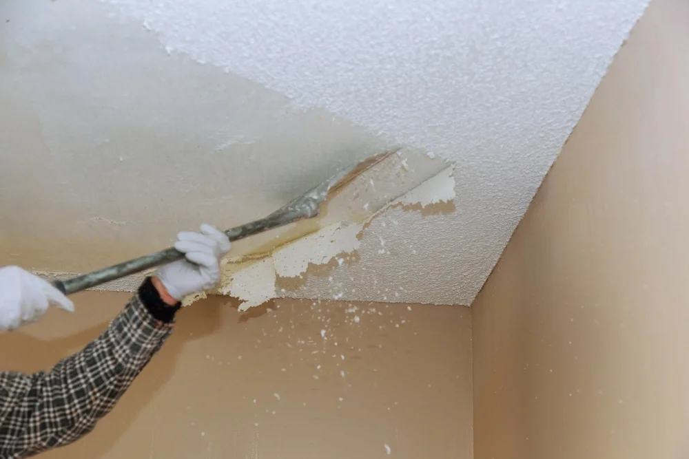 Worker scraping a white popcorn ceiling with a long-handled tool